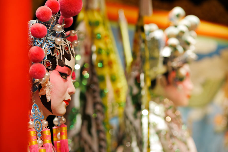 Primer plano de perfil de un maniquí o actriz de la ópera china tradicional con maquillaje teatral detallado y un elaborado tocado con pompones rojos y perlas, con otra figura desenfocada al fondo.