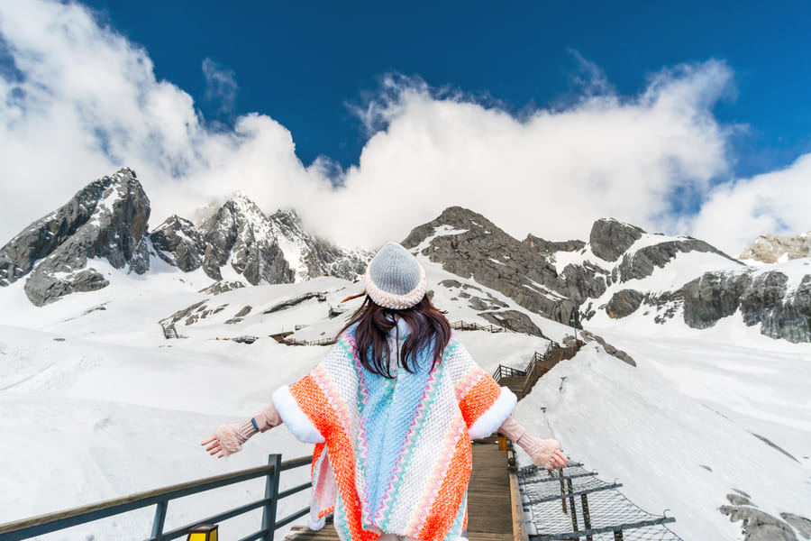 Mujer de pie en un puente en frente de una montaña con nieve, vistinedo una capa blanca con detalles azules y naranjas y un gorro.